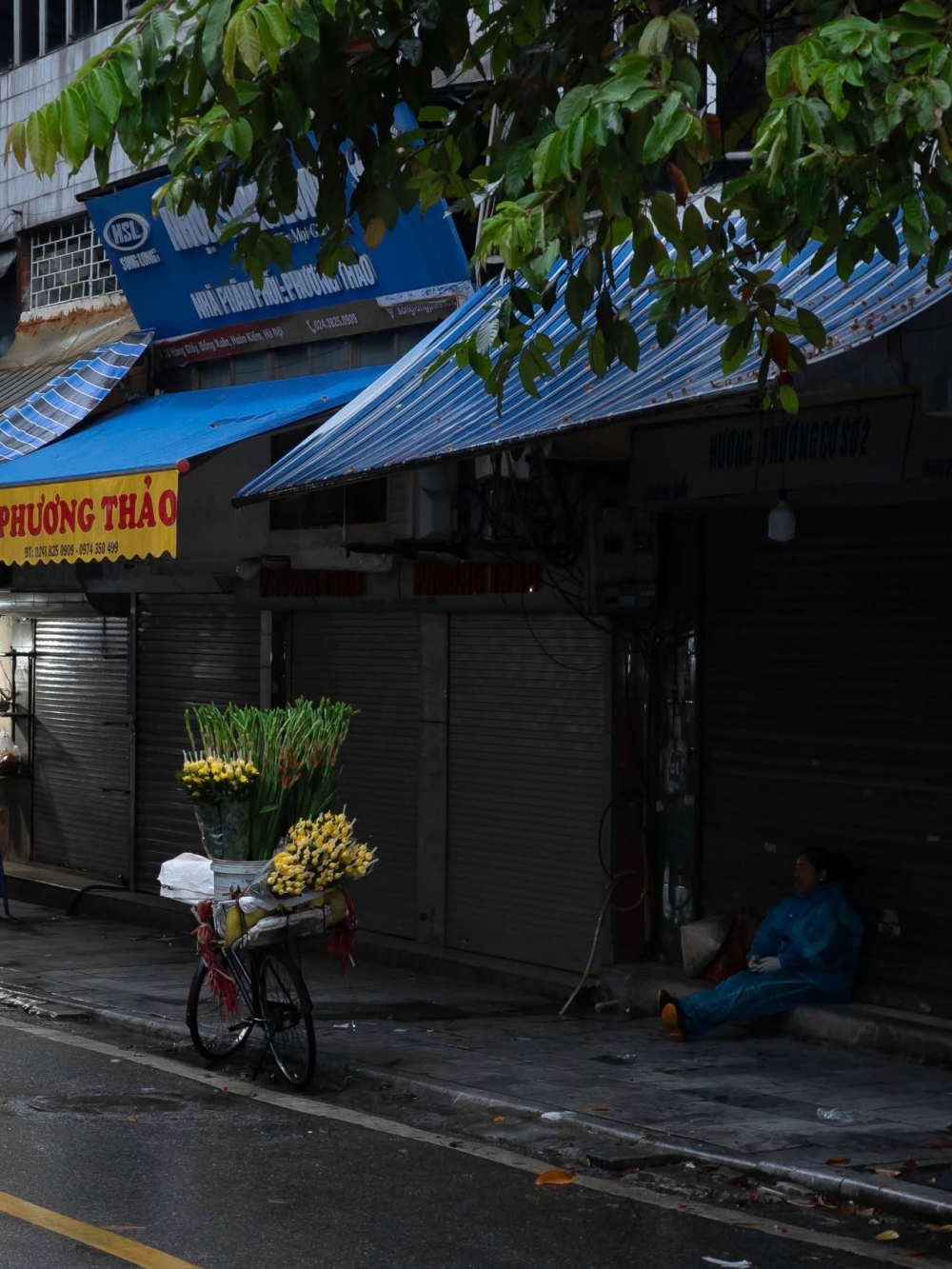 Giữa tiết trời mưa lạnh, người bán hàng rong tạm nghỉ bên gánh hoa bên vỉa hè, tìm chút hơi ấm giữa nhịp sống Hà Nội đầu đông